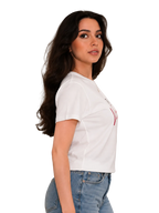 A woman with long dark hair is shown in profile wearing a fitted white short-sleeve T-shirt with a small red graphic on the chest and light blue jeans, standing against a plain light beige wall.