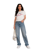 A woman stands in a studio setting wearing a white Valentino graphic T-shirt, light-wash wide-leg jeans, white sneakers, and holding a small white handbag.