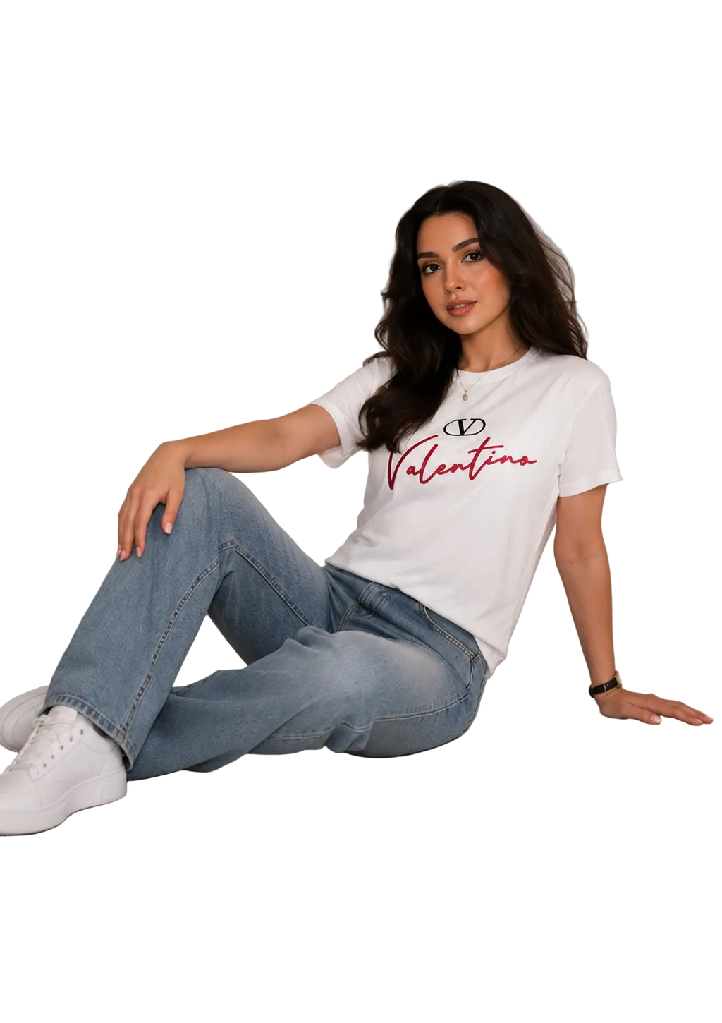A woman sitting on a light gray floor against a white paneled wall, wearing a white Valentino graphic t-shirt, light blue jeans, and white sneakers.