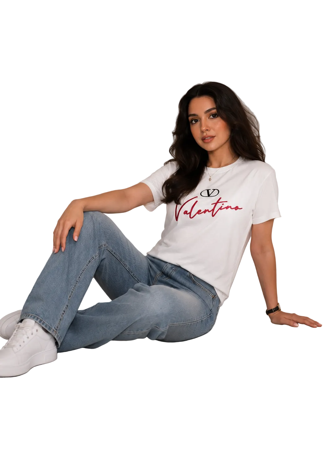 A woman sitting on a light gray floor against a white paneled wall, wearing a white Valentino graphic t-shirt, light blue jeans, and white sneakers.