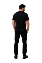 A man viewed from behind wearing a black short-sleeve t-shirt and black pants, standing against a neutral gray studio background with soft shadows.