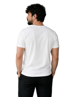 A man viewed from behind wearing a plain white short-sleeve crewneck t-shirt and black pants, standing in a minimalist indoor setting with a gray wall and a potted plant.