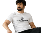 A man with a beard and curly hair sits on a beige sofa wearing a white Balenciaga Paris t-shirt with a black printed logo and text.