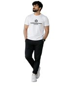 A man wearing a white Balenciaga t-shirt with black joggers and white sneakers, walking in a modern indoor setting with a glass wall and staircase.