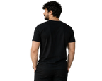 A man viewed from behind wearing a plain black short-sleeve crewneck t-shirt and black pants, standing in a modern indoor setting with a gray wall and a potted plant.