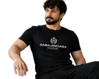A man with a beard and curly hair sits on a light beige sofa wearing a black Balenciaga Paris t-shirt with a white printed logo and text.
