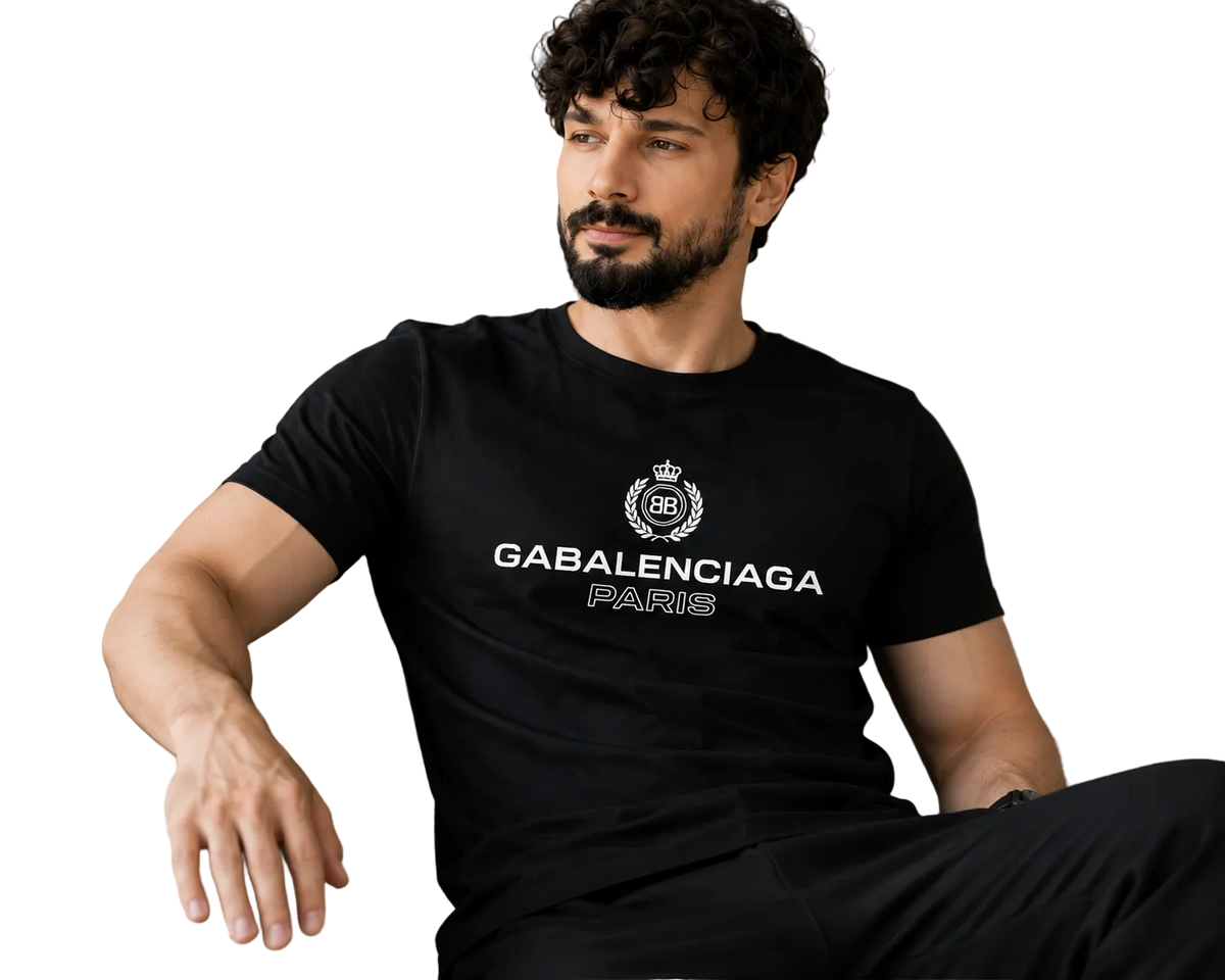 A man with a beard and curly hair sits on a light beige sofa wearing a black Balenciaga Paris t-shirt with a white printed logo and text.