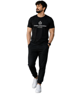 A man wearing a black Balenciaga short-sleeve t-shirt with a white logo, black jogger pants, and white sneakers, standing in a modern indoor setting with a glass door and staircase.