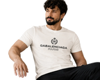 A man with a beard and curly hair sits on a beige couch wearing a white t-shirt with a black GABALENCIAGA logo and text on the chest.