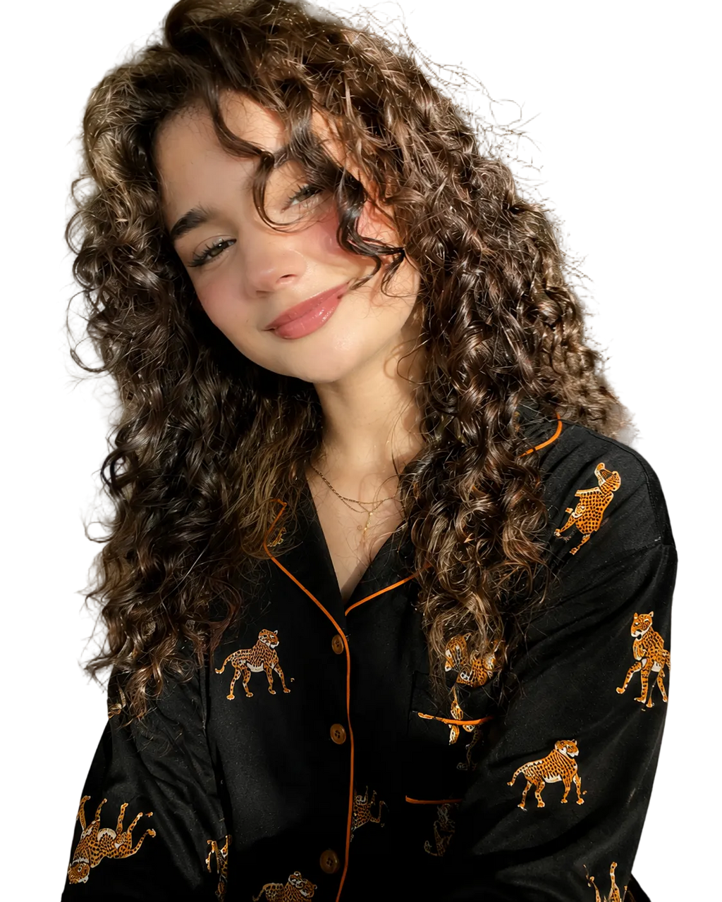 A woman with long, curly brown hair wearing a black leopard print pajama set with orange piping, posing in a bedroom setting with warm sunlight.