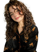 A woman with long, curly brown hair wearing a black leopard print pajama set with orange piping, posing in a bedroom setting with warm sunlight.