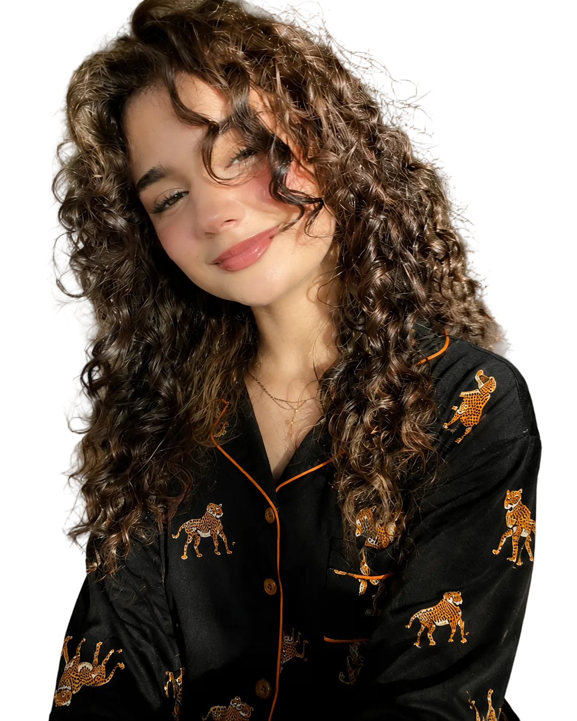 A woman with long, curly brown hair wearing a black leopard print pajama set with orange piping, posing in a bedroom setting with warm sunlight.