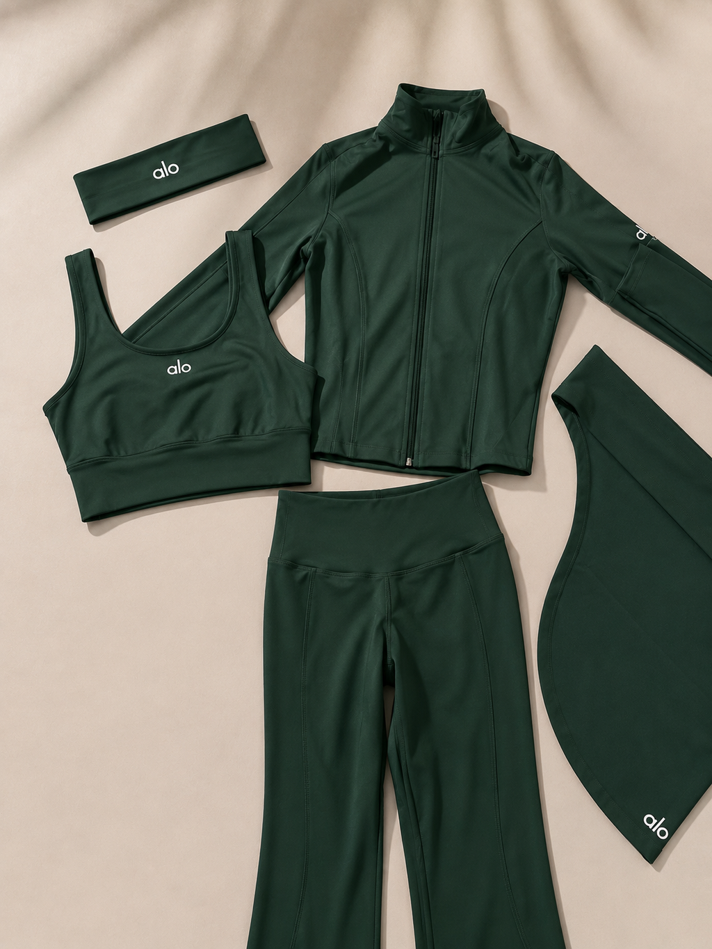 A dark green activewear set laid out on a beige fabric background, including a zip-front jacket, sports bra, leggings, headband, and a curved sleeve or wrist cover. Each piece has a small white 'alo' logo.