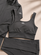 A dark gray three-piece activewear set from alo, featuring a sports bra, high-waisted leggings, and a zip-up jacket, laid flat on a beige background with soft shadows.