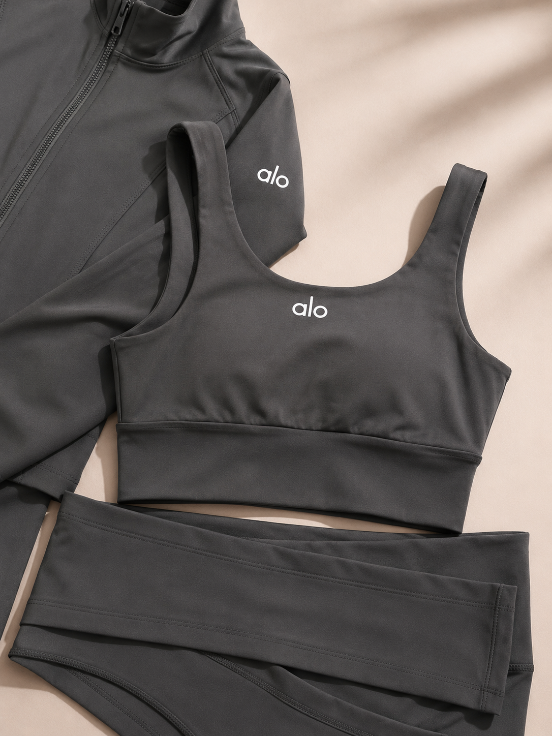 A dark gray three-piece activewear set from alo, featuring a sports bra, high-waisted leggings, and a zip-up jacket, laid flat on a beige background with soft shadows.
