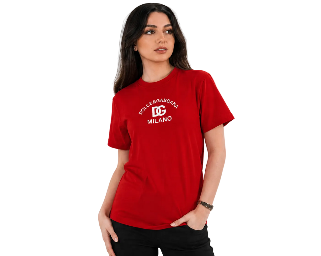 A woman modeling a red short-sleeve t-shirt with a white printed Dolce & Gabbana Milano logo on the chest, set against a plain light gray background.