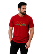 A man wearing a red short-sleeve t-shirt with a yellow and green Gucci logo printed across the chest, paired with black pants, set against a plain gray studio background.