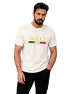 A man wearing a cream-colored Gucci t-shirt with a yellow logo and green and red stripe detail, paired with black pants, against a solid gray studio background.