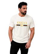 A man wearing a cream-colored Gucci t-shirt with a yellow logo and the classic green and red web stripe, paired with black pants, against a neutral gray background.