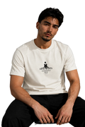 A man wearing a white crew-neck t-shirt with a black graphic print on the chest, paired with black pants, sitting in a modern room with a gray wall and a black leather sofa in the background.