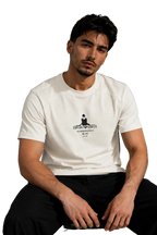 A man wearing a white crew-neck t-shirt with a black graphic print on the chest, paired with black pants, sitting in a modern room with a gray wall and a black leather sofa in the background.