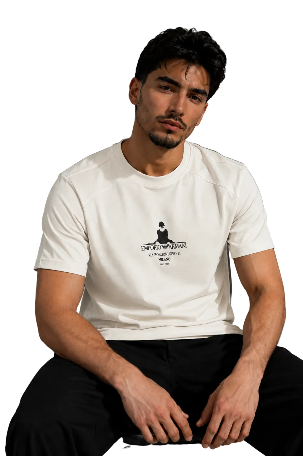 A man wearing a white crew-neck t-shirt with a black graphic print on the chest, paired with black pants, sitting in a modern room with a gray wall and a black leather sofa in the background.
