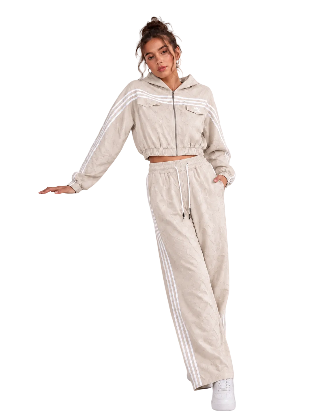 A woman models a matching beige cropped zip-up hoodie and wide-leg sweatpants set with white side stripes, posing next to a white display block in a studio setting.