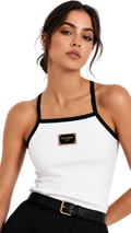 A woman wearing a white ribbed knit halter top with black trim and a gold-tone rectangular brand patch on the chest, paired with black pants and a black belt against a light gray studio background.