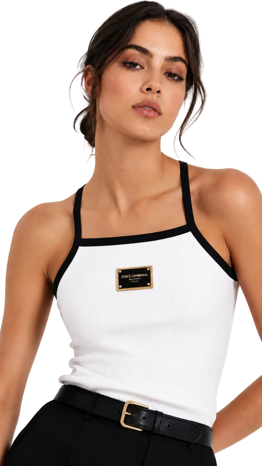 A woman wearing a white ribbed knit halter top with black trim and a gold-tone rectangular brand patch on the chest, paired with black pants and a black belt against a light gray studio background.