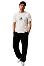 A man wearing a white crew-neck t-shirt with a black graphic print on the chest, paired with black pants and white sneakers, standing in a modern gray room.