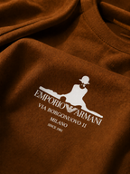A close-up of a brown crewneck sweatshirt featuring a white printed Emporio Armani logo and text on the chest.