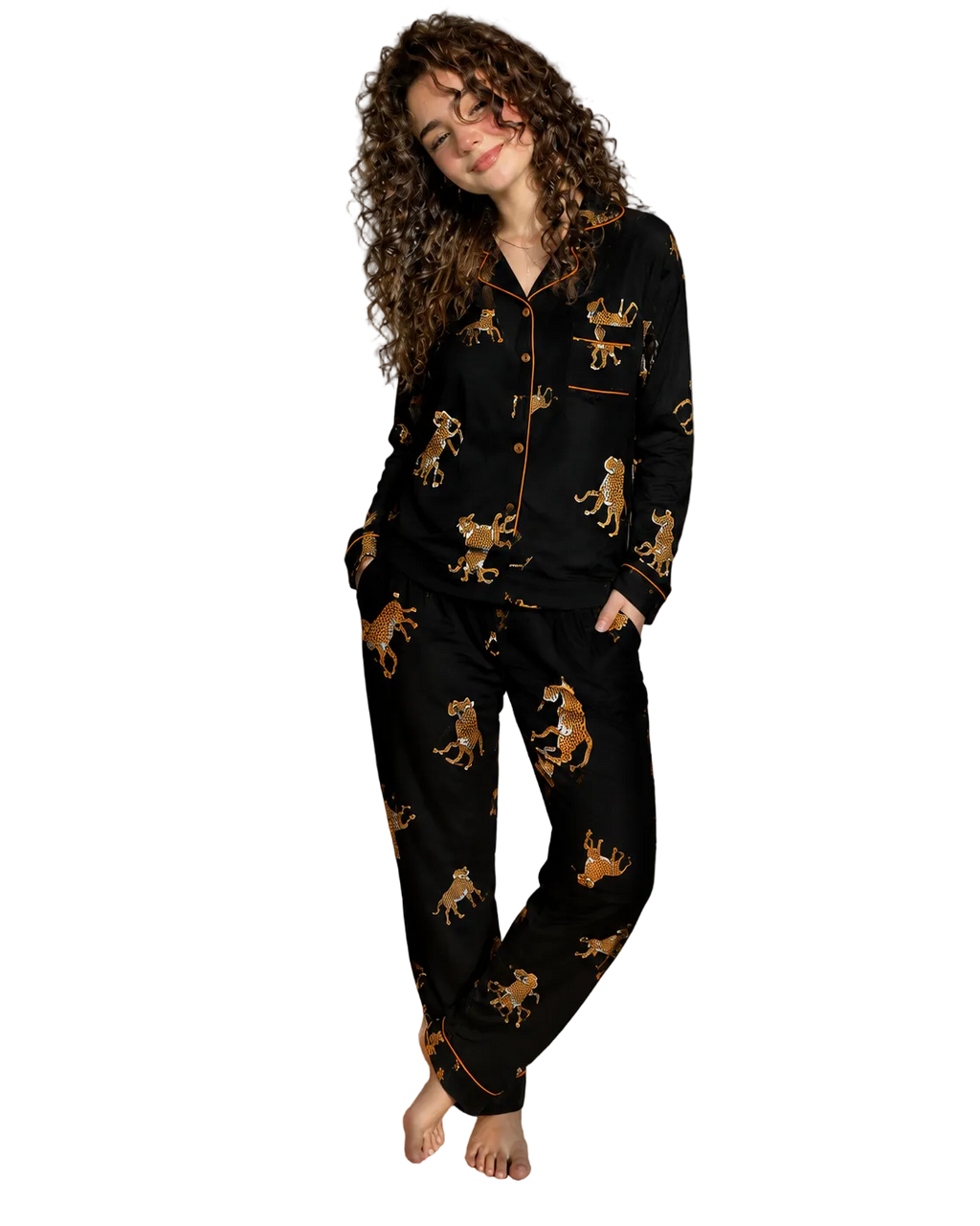 A woman stands barefoot in a bedroom wearing a black pajama set with a gold and brown leopard print and gold piping. The set includes a long-sleeve button-front top and matching pants.