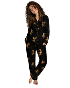 A woman stands barefoot in a bedroom wearing a black pajama set with a gold and brown leopard print and gold piping. The set includes a long-sleeve button-front top and matching pants.
