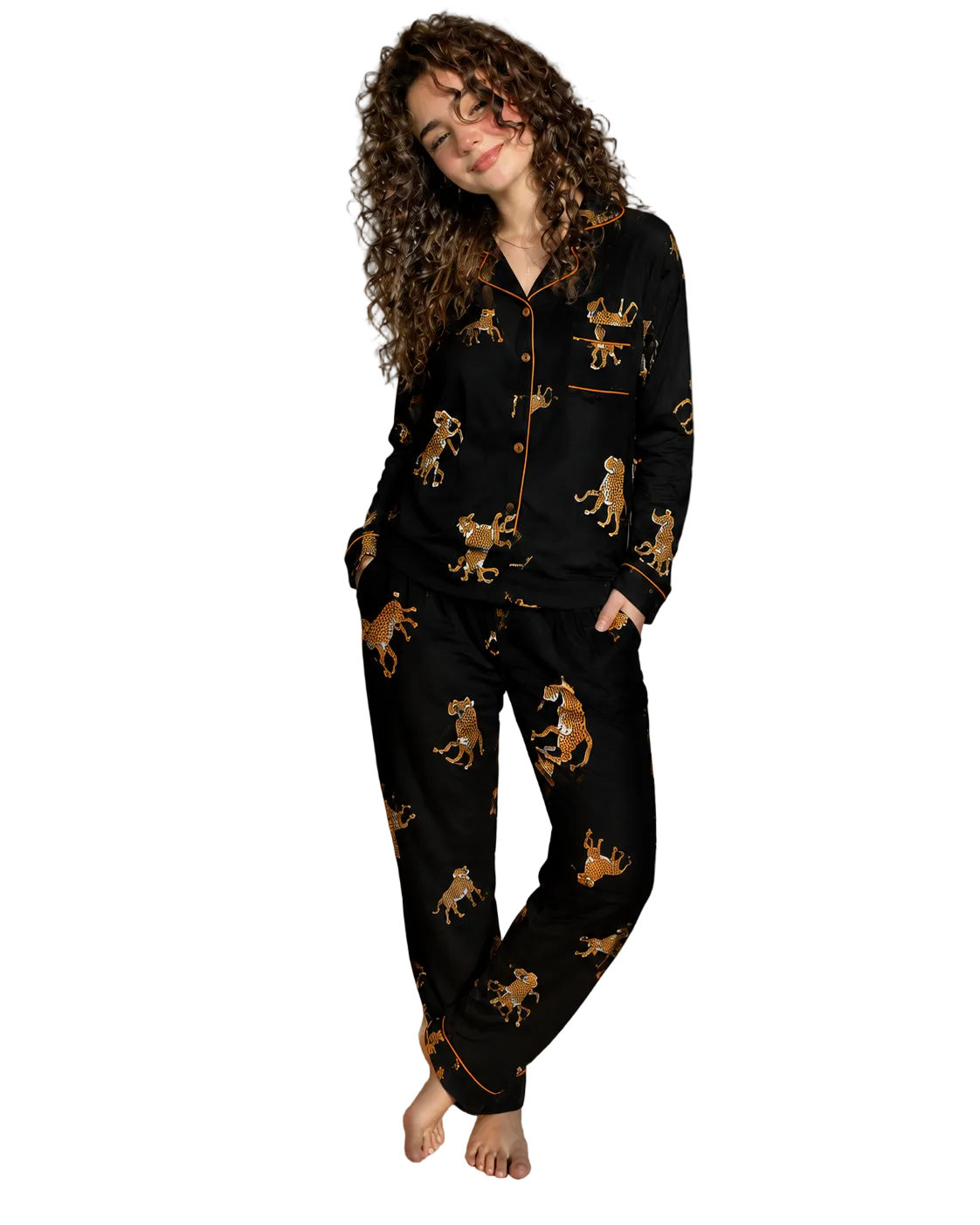 A woman stands barefoot in a bedroom wearing a black pajama set with a gold and brown leopard print and gold piping. The set includes a long-sleeve button-front top and matching pants.