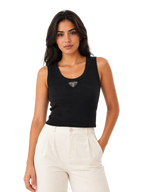 A woman modeling a black ribbed knit sleeveless tank top with a small silver-toned triangular logo detail at the center of the chest, paired with cream-colored pants, against a plain white studio background.