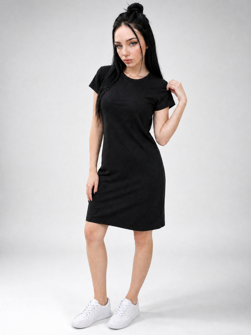 A woman modeling a plain black short-sleeve t-shirt dress with side pockets, paired with white sneakers, against a light gray studio background.