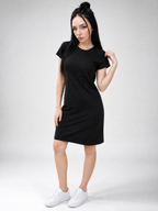 A woman modeling a plain black short-sleeve t-shirt dress with side pockets, paired with white sneakers, against a light gray studio background.