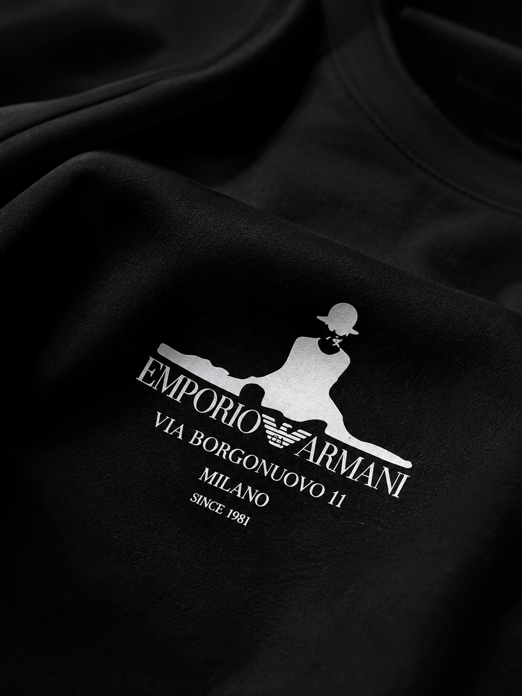 Close-up of a black crewneck sweatshirt featuring a white screen-printed graphic of the Emporio Armani logo and text on the chest.