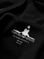 Close-up of a black crewneck sweatshirt featuring a white screen-printed graphic of the Emporio Armani logo and text on the chest.