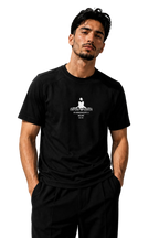 A man wearing a black short-sleeve t-shirt with a white graphic print on the chest, paired with black pants, standing against a textured gray wall.