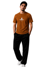 A man wearing a rust-orange crewneck t-shirt with a white graphic print on the chest, paired with black pants and white sneakers, standing in a modern gray room.