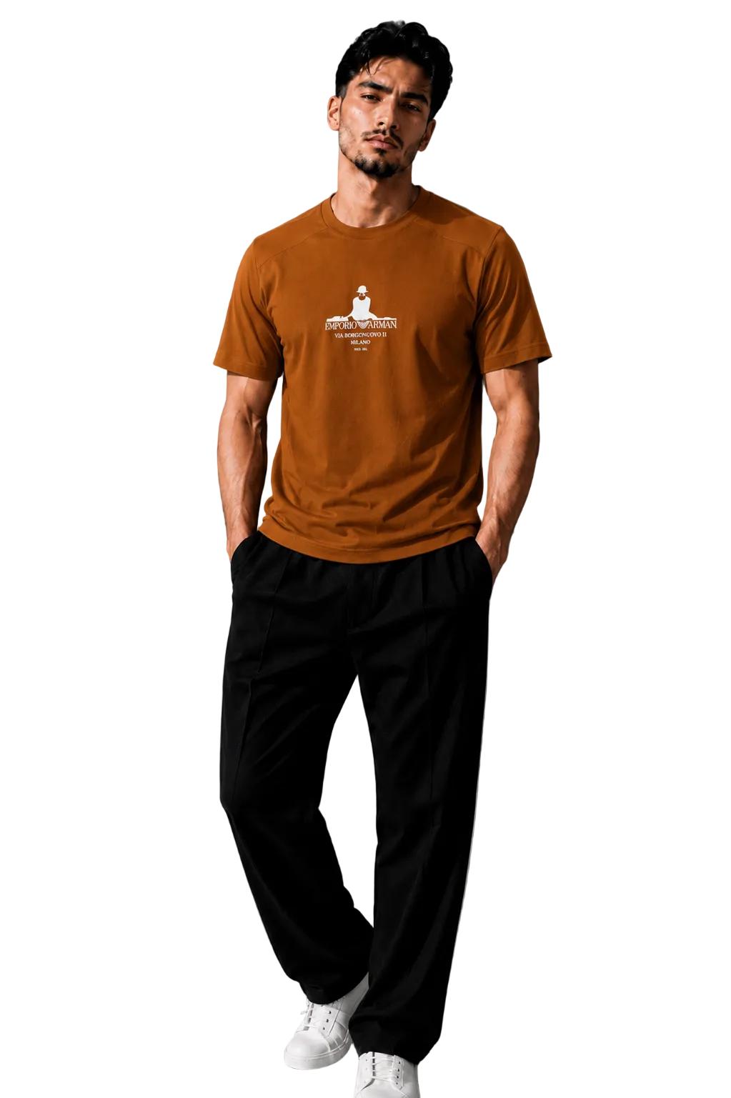 A man wearing a rust-orange crewneck t-shirt with a white graphic print on the chest, paired with black pants and white sneakers, standing in a modern gray room.