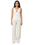 A woman modeling a white sleeveless tank top with a small black diamond-shaped logo on the chest, paired with light beige wide-leg trousers and white sneakers, against a plain white background.