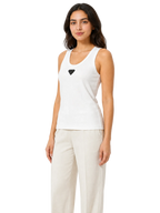 A woman modeling a white sleeveless tank top with a small black triangular logo on the chest, paired with light beige pants, against a plain white background.