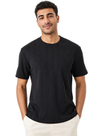 A man wearing a black short-sleeve ribbed crewneck t-shirt and off-white pants, standing against a solid white background.