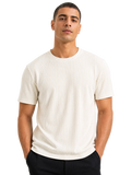 A man wearing a ribbed short-sleeve crewneck t-shirt in off-white, paired with black pants, photographed against a solid white background.