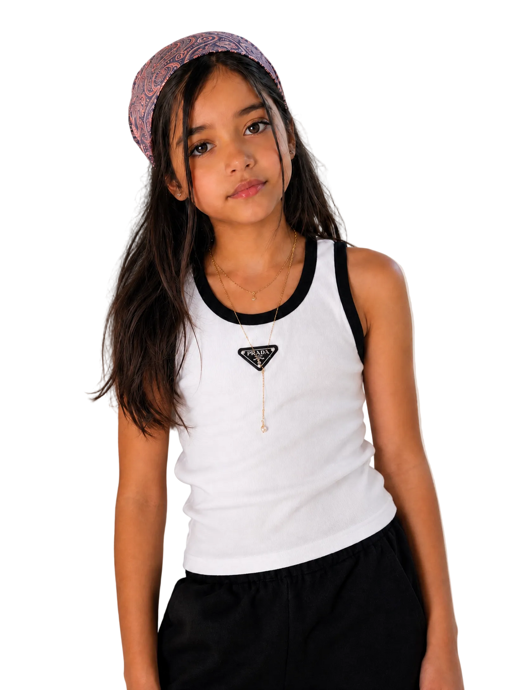A young girl with long dark hair wearing a white tank top with black trim, a black Prada triangle necklace, and a patterned headband, posing against a plain light beige wall.