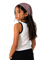 A young girl with long dark hair is shown from the back and side, wearing a white ribbed tank top with black trim, black pants, and a patterned mauve and blue bandana headwrap against a plain light beige studio background.