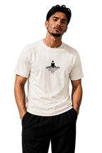 A man wearing a white crew-neck t-shirt with a black graphic print on the chest, paired with black pants, standing against a textured gray wall.