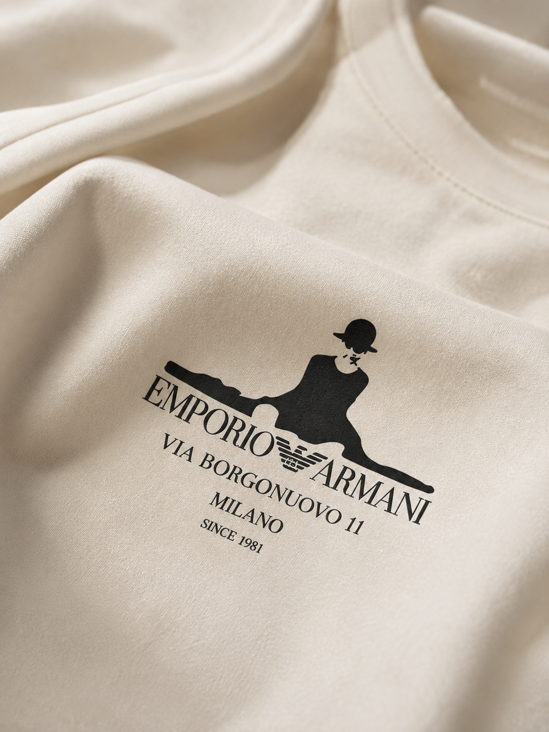 Close-up of an off-white crewneck sweatshirt featuring a black printed graphic of a seated figure and the text 'EMPORIO ARMANI VIA BORGONUOVO II MILANO SINCE 1981'.
